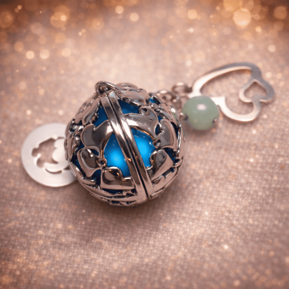 bola de grossesse personnalise Bola de grossesse en acier argenté au design ajouré, sublimé par une pierre bleue lumineuse. Son tintement doux favorise la détente et le bien-être pendant la grossesse. Accompagné de charms délicats (trèfle, cœur ou perle), ce bijou symbolise la protection, la sérénité et l’harmonie. Idéal comme cadeau de grossesse ou bijou de maternité.